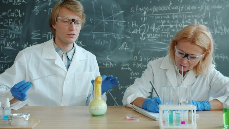 Scientist wearing blue gloves prepares a vaccine dose by drawing liquid from a vial using a syringe under a sterile laboratory hood, with test tubes and medical supplies visible in the background.